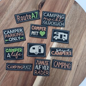 Könnte beinhalten: Eine Sammlung schwarz-weißer gestickter Aufnäher mit Camping-Motiven. Die Aufnäher enthalten Sätze wie "Route A7", "Camping macht glücklich" und "Camper Life". Die Aufnäher sind auf einer Holzoberfläche angeordnet.