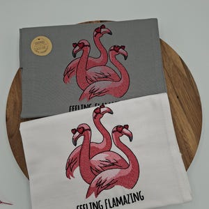Könnte beinhalten: Zwei bestickte Tücher mit Flamingo-Motiv. Eines ist grau, das andere weiß. Jedes zeigt drei rosa Flamingos mit herzförmigen Sonnenbrillen. Der Schriftzug "FEELING FLAMAZING" ist auf das weiße Tuch gestickt.