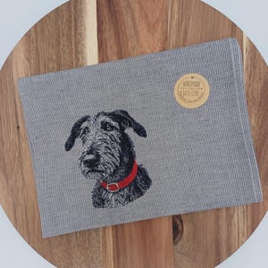 Könnte beinhalten: Ein grau-weiß gemusterter Stoff mit einem gestickten Hundeporträt mit rotem Halsband. Ein runder Aufkleber mit dem Text "HANDMADE WITH LOVE" befindet sich auf dem Stoff. Der Stoff liegt auf einem Holzschneidebrett.