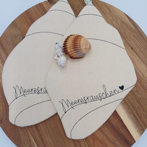 Könnte beinhalten: Zwei beige Topflappen in Muschelform mit schwarzen Nähten und dem Schriftzug "Meeresrauschen". Auf den Topflappen liegen eine braun-weiße Muschel und eine kleine weiße Muschel. Die Topflappen haben Aufhängeschlaufen mit grün-weißem Muster.