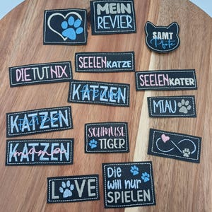 Könnte beinhalten: Eine Sammlung schwarzer, gestickter Aufnäher mit weißen Nähten. Die Aufnäher zeigen verschiedene Texte und Designs, darunter Pfotenabdrücke, Herzen und das Wort "LOVE". Einige Aufnäher haben deutschen Text.