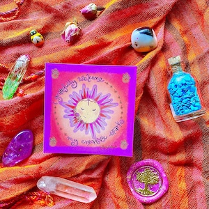 May include: A pink card with a sun illustration and the text "I gratefully welcome my creative spark". The card is surrounded by crystals, a small bottle of blue glitter, and other decorative items on a red and orange fabric.