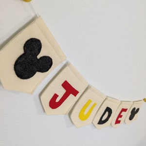 Pode incluir: Uma faixa de feltro com o nome "JUDE" escrito em letras de feltro vermelho, amarelo, preto e vermelho. A faixa &eacute; decorada com uma cabe&ccedil;a de Mickey Mouse de feltro preto em cada extremidade.