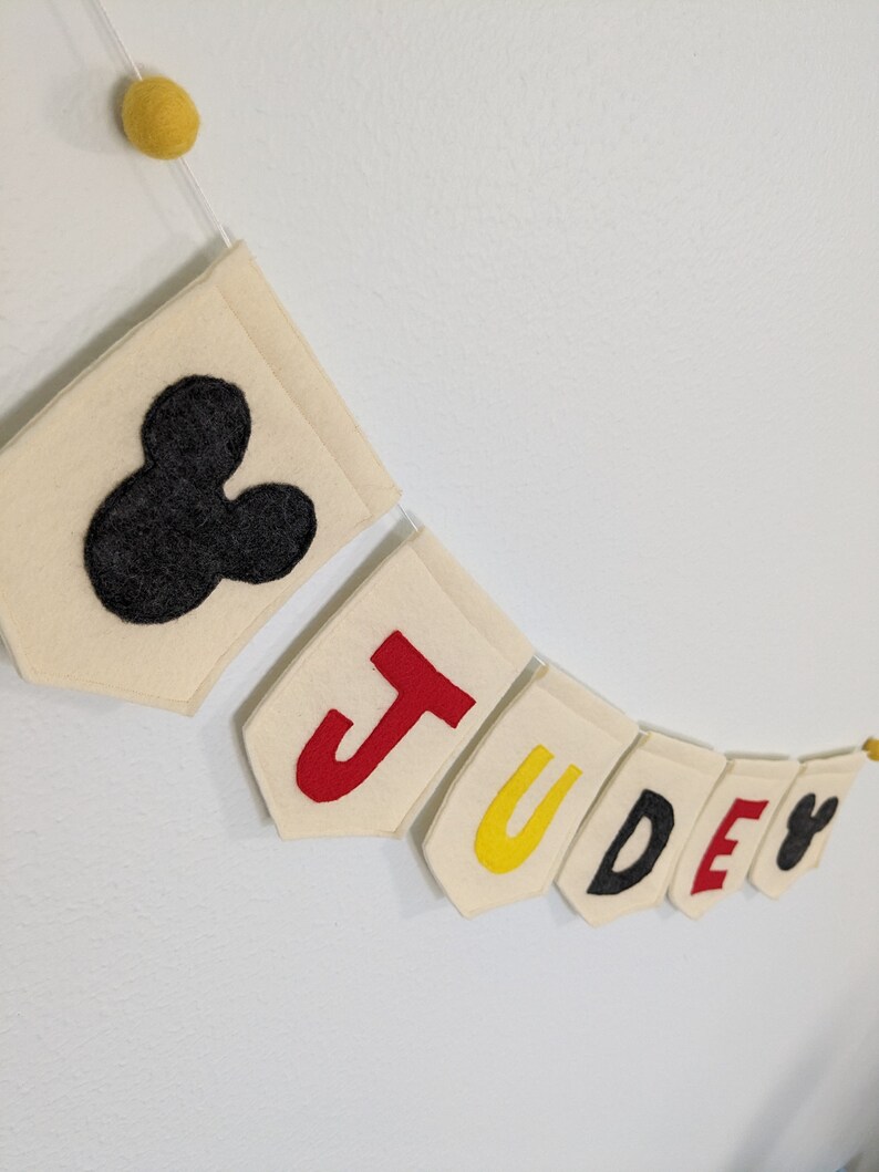 Pode incluir: Uma faixa de feltro com o nome "JUDE" escrito em letras de feltro vermelho, amarelo, preto e vermelho. A faixa &eacute; decorada com uma cabe&ccedil;a de Mickey Mouse de feltro preto em cada extremidade.