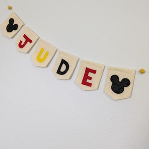 Pode incluir: Uma faixa de feltro branco com o nome "JUDE" escrito em letras de feltro vermelho, amarelo e preto. A faixa &eacute; decorada com duas orelhas de Mickey Mouse de feltro preto em cada extremidade.