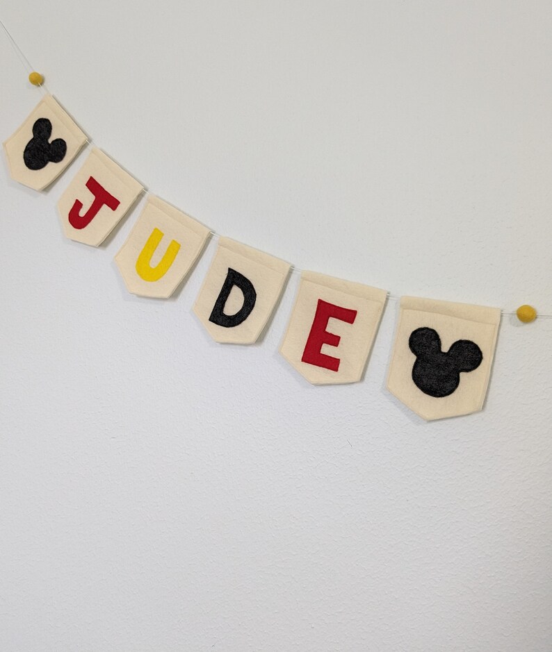 Pode incluir: Uma faixa de feltro branco com o nome "JUDE" escrito em letras de feltro vermelho, amarelo e preto. A faixa &eacute; decorada com duas orelhas de Mickey Mouse de feltro preto em cada extremidade.