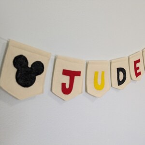 Pode incluir: Uma faixa de feltro branco com o nome "JUDE" escrito em letras de feltro vermelho, amarelo e preto. Cada letra est&aacute; em uma bandeira separada. A faixa &eacute; decorada com orelhas de Mickey Mouse de feltro preto nas pontas.