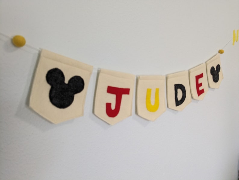 Pode incluir: Uma faixa de feltro branco com o nome "JUDE" escrito em letras de feltro vermelho, amarelo e preto. Cada letra est&aacute; em uma bandeira separada. A faixa &eacute; decorada com orelhas de Mickey Mouse de feltro preto nas pontas.