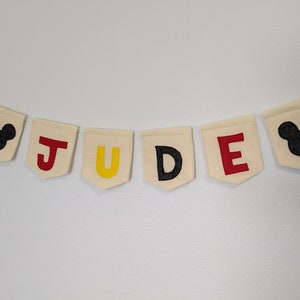 Pode incluir: Uma faixa de feltro branco com o nome "JUDE" escrito em letras de feltro vermelho, amarelo e preto. Cada letra est&aacute; em uma bandeira separada. A faixa &eacute; decorada com duas cabe&ccedil;as de Mickey Mouse de feltro preto nas pontas.