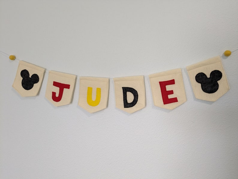 Pode incluir: Uma faixa de feltro branco com o nome "JUDE" escrito em letras de feltro vermelho, amarelo e preto. Cada letra est&aacute; em uma bandeira separada. A faixa &eacute; decorada com duas cabe&ccedil;as de Mickey Mouse de feltro preto nas pontas.