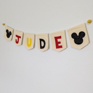 Pode incluir: Uma faixa de feltro com o nome "JUDE" em letras vermelhas, amarelas e pretas. A faixa &eacute; decorada com duas orelhas de Mickey Mouse de feltro preto em cada extremidade.