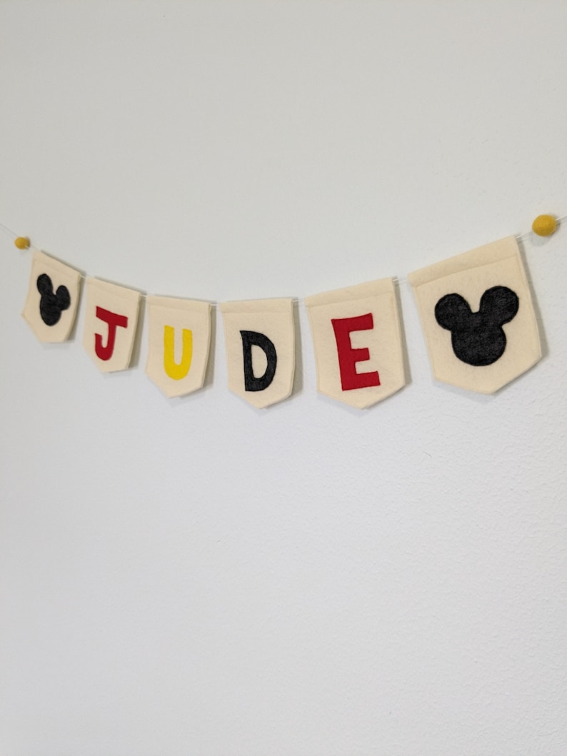 Pode incluir: Uma faixa de feltro com o nome "JUDE" em letras vermelhas, amarelas e pretas. A faixa &eacute; decorada com duas orelhas de Mickey Mouse de feltro preto em cada extremidade.