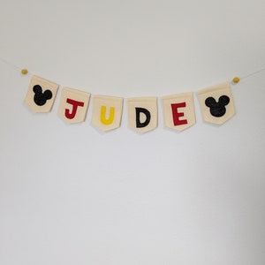 Pode incluir: Uma faixa de tecido branco com o nome "JUDE" escrito em letras de feltro vermelho, amarelo e preto. A faixa &eacute; decorada com orelhas de Mickey Mouse de feltro preto em cada extremidade.