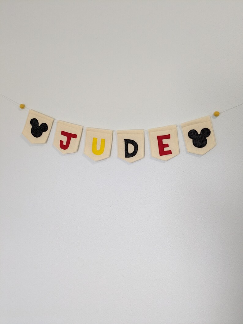 Pode incluir: Uma faixa de tecido branco com o nome "JUDE" escrito em letras de feltro vermelho, amarelo e preto. A faixa &eacute; decorada com orelhas de Mickey Mouse de feltro preto em cada extremidade.