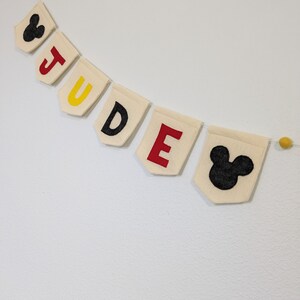 Pode incluir: Uma faixa de feltro com o nome "JUDGE" escrito em letras pretas em bandeiras de feltro branco. Cada letra &eacute; acentuada com uma cor diferente: amarelo, vermelho e amarelo. A faixa &eacute; decorada com duas cabe&ccedil;as pretas do Mickey Mouse nas pontas.