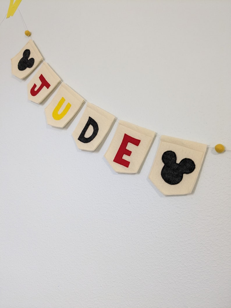 Pode incluir: Uma faixa de feltro com o nome "JUDGE" escrito em letras pretas em bandeiras de feltro branco. Cada letra &eacute; acentuada com uma cor diferente: amarelo, vermelho e amarelo. A faixa &eacute; decorada com duas cabe&ccedil;as pretas do Mickey Mouse nas pontas.
