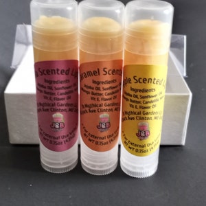 May include: Three lip balm tubes with different scents: Apple, Caramel, and a third scent that is not visible. Each tube is labeled with the ingredients, which include jojoba oil, sunflower oil, mango butter, candelilla wax, vitamin E, flavor oil, and the company name "Mythical Gardens". The tubes are 0.15 oz (4.25 g) each.