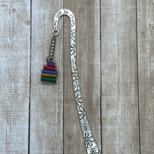 May include: A silver metal bookmark with a floral design and a stack of colorful books charm. The bookmark is curved at the top and has a decorative pattern of flowers and other shapes.