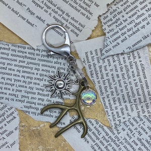 May include: A keychain with a silver clasp, featuring a sun charm, a bronze antler charm, and a shimmering iridescent charm. The background includes torn paper with printed text.