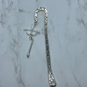 May include: Silver-colored metal bookmark featuring a decorative design. The bookmark has a curved top and a dangling charm of a bow and arrow. The bookmark's design includes a detailed pattern along the stem and a foot-like shape at the bottom.