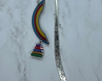 May include: A silver metal bookmark with a rainbow and stack of books charm. The rainbow is blue, red, yellow, and orange. The books are various colors. The bookmark has a floral design.
