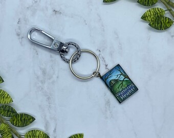 May include: A silver-toned keychain with a rectangular charm featuring a miniature painting of a house on a hill under a night sky. The charm reads "Wuthering Heights" in white text. The keychain has a clasp and a ring.