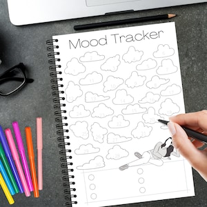 May include: A spiral-bound notebook with the words "Mood Tracker" at the top. The notebook features cloud shapes numbered 1-30 for tracking moods. A person is shown drawing in the notebook with a black pen. Colorful markers and a cup of coffee are also visible.