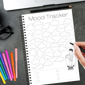 Puede incluir: Un cuaderno de espiral con un diseño de "Mood Tracker". El rastreador presenta formas de nubes numeradas del 1 al 31, con una pequeña ilustración de una persona. Rotuladores de colores, una taza de café y gafas están sobre la superficie gris oscura.