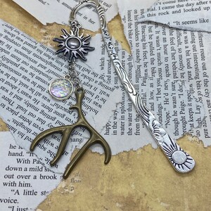 Peut inclure: Marque-page en métal avec un motif de soleil et un charm en forme de soleil, un charm irisé en forme d'écailles et un charm en forme de bois de cerf en bronze. Le marque-page est posé sur un fond de papier déchiré.