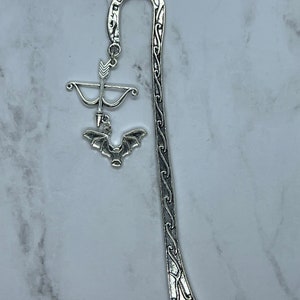 May include: Silver-toned metal bookmark featuring a decorative design. The bookmark has a curved top and a long, slender body with an ornate pattern. A charm with a bow and arrow and a bat hangs from the top.