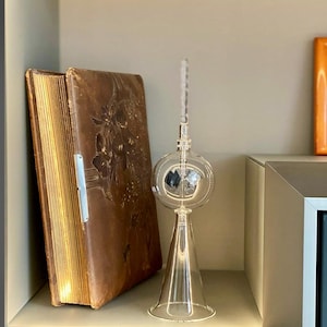 Könnte beinhalten: Ein Vintage-Buch mit Lederbindung und goldenen Akzenten liegt auf einem Regal neben einem durchsichtigen Glasobjekt mit einer runden Kugel und einer konischen Basis.