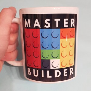 May include: White ceramic mug with a black background and the words "MASTER BUILDER" in white text. The background features a colorful design of nine red, blue, green, and yellow Lego bricks.