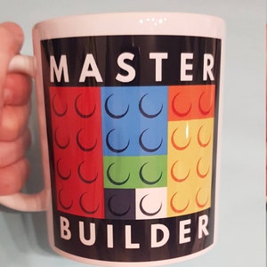 May include: White ceramic mug with a black background and the text "MASTER BUILDER" in white letters. The background features a colorful design of red, blue, green, yellow, black, and white Lego bricks.