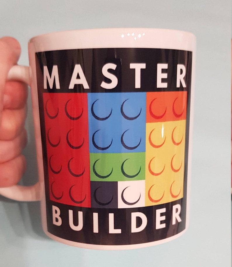 May include: White ceramic mug with a black background and the text "MASTER BUILDER" in white letters. The background features a colorful design of red, blue, green, yellow, black, and white Lego bricks.