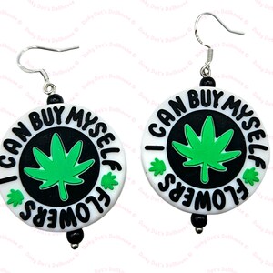 Puede incluir: Un par de pendientes redondos blancos y negros con el texto "I CAN BUY MYSELF FLOWERS" en un diseño circular. Cada pendiente presenta una hoja de cannabis verde en el centro. Los pendientes tienen ganchos plateados y cuentas negras.