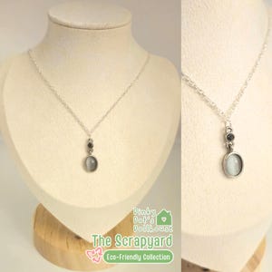 May include: A silver necklace with a pendant featuring a gray oval stone and two smaller dark stones. The necklace is displayed on a mannequin. The text "Dinky Dot's Dollhouse" and "The Scrapyard" are visible.