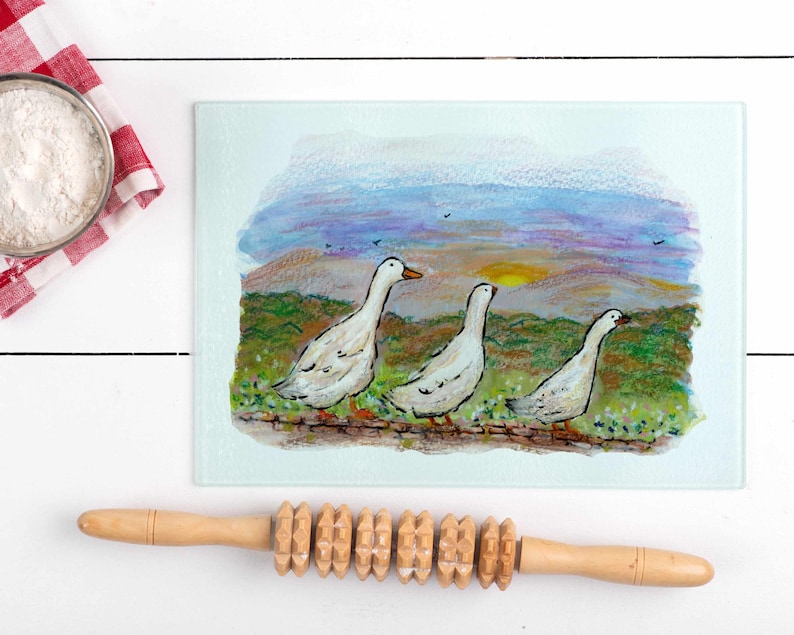 May include: A rectangular glass cutting board with a painted scene of three white geese walking along a grassy area with a sunset background. A wooden rolling pin is below the board. A bowl of flour sits on a red and white checkered cloth.