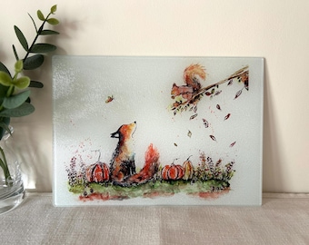 May include: A rectangular glass cutting board featuring a watercolor illustration of a fox sitting among orange pumpkins and green foliage, looking up at a squirrel perched on a tree branch with falling leaves and a small butterfly in the air.