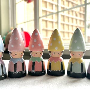 May include: Six hand-painted wooden gnome figurines. Each gnome has a conical hat with white polka dots, and a painted face. The gnomes wear colourful outfits in shades of blue, pink, yellow, and green. The figurines are approximately 10 cm tall.