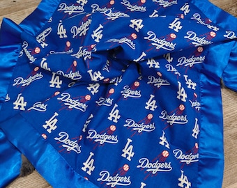 Puede incluir: Una manta azul para bebé con borde de satén. La manta tiene el logotipo de los Dodgers de Los Ángeles y pelotas de béisbol. El nombre y el logotipo del equipo son blancos, con detalles en rojo.