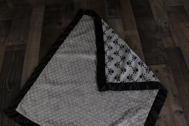 May include: A gray and black baby blanket with a skull and crossbones pattern on one side and a soft, textured fabric on the other side.