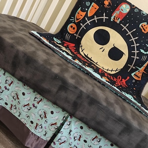 Puede incluir: Una manta gris oscuro con una manta negra con un dise&ntilde;o de Jack Skellington. Tambi&eacute;n se ve una manta azul claro con un patr&oacute;n repetido de Jack Skellington y Sally. Las mantas est&aacute;n sobre una cama.
