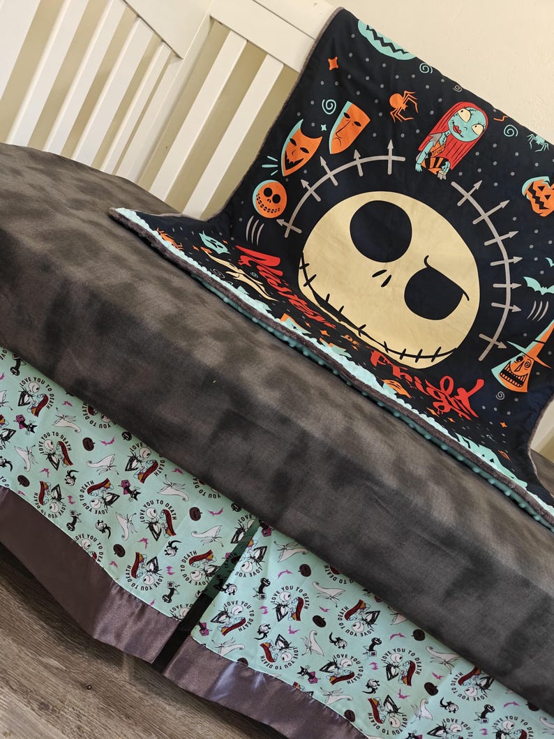 Puede incluir: Una manta gris oscuro con una manta negra con un dise&ntilde;o de Jack Skellington. Tambi&eacute;n se ve una manta azul claro con un patr&oacute;n repetido de Jack Skellington y Sally. Las mantas est&aacute;n sobre una cama.