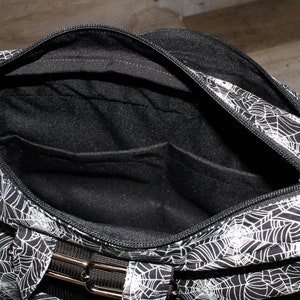 May include: Black and white spiderweb patterned bag with a zipper closure and a black strap. The bag has a black interior with a zippered pocket.
