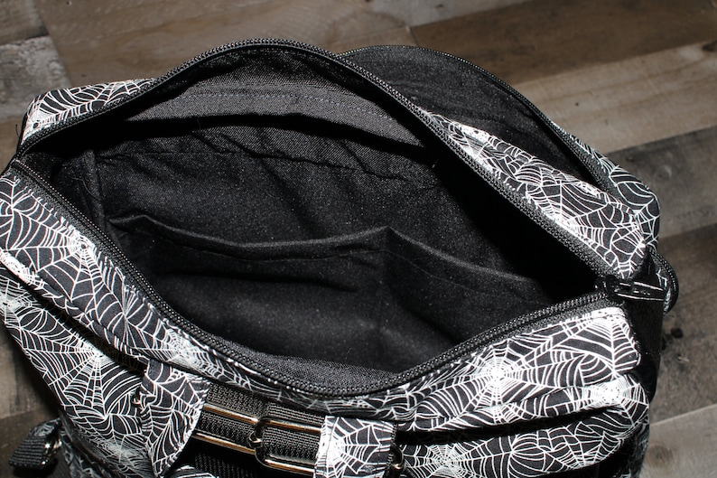 May include: Black and white spiderweb patterned bag with a zipper closure and a black strap. The bag has a black interior with a zippered pocket.