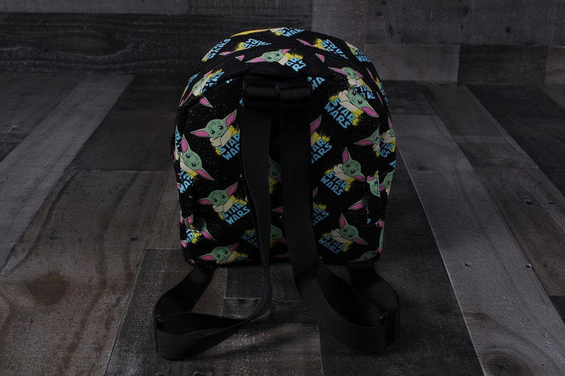 Puede incluir: Mochila negra con un patr&oacute;n repetido de un personaje verde y azul con las palabras "Star Wars" sobre un fondo negro.