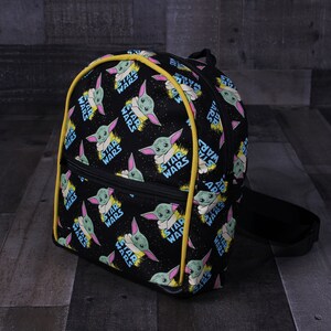 Puede incluir: Mochila negra con ribete amarillo. La mochila est&aacute; cubierta con un patr&oacute;n repetido de un personaje alien&iacute;gena verde con las palabras "Star Wars" impresas en ella.