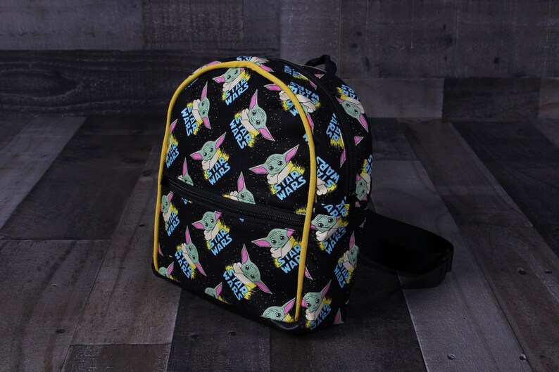 Puede incluir: Mochila negra con ribete amarillo. La mochila est&aacute; cubierta con un patr&oacute;n repetido de un personaje alien&iacute;gena verde con las palabras "Star Wars" impresas en ella.