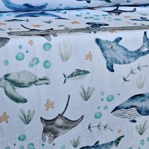 May include: White fabric with a watercolor-style ocean pattern. The design features dolphins, fish, sea turtles, stingrays, jellyfish, and whales in shades of blue, green, and gray, with accents of orange and teal. The fabric is rolled up in the background.
