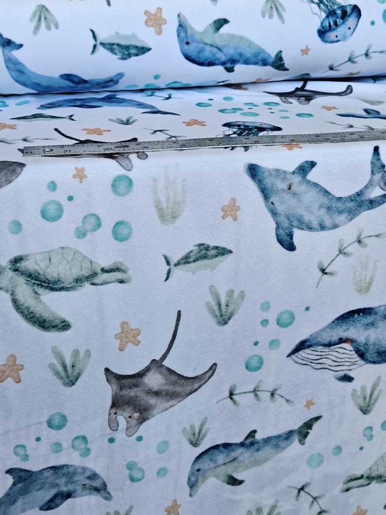May include: White fabric with a watercolor-style ocean pattern. The design features dolphins, fish, sea turtles, stingrays, jellyfish, and whales in shades of blue, green, and gray, with accents of orange and teal. The fabric is rolled up in the background.